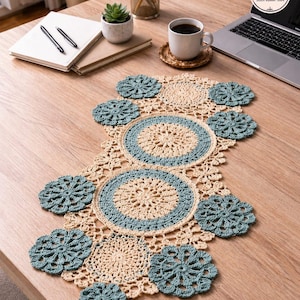 May include: A beige and teal crochet table runner with matching coasters. The runner has a central design of concentric circles and floral patterns. The coasters are teal and flower-shaped. The runner is displayed on a wooden table.