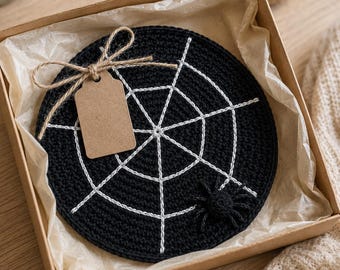 May include: A round, black crocheted coaster featuring a white spiderweb pattern and a small, attached black spider. The coaster is presented in a square cardboard gift box lined with light-colored tissue paper and includes a blank brown paper tag.