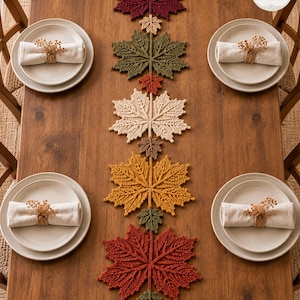 May include: A crochet table runner featuring a vertical arrangement of maple leaves in shades of burgundy, olive green, cream, mustard yellow, and rust, accented by smaller green and brown leaves, placed on a wooden table with four place settings.