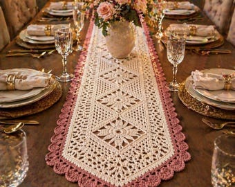 May include: A rectangular crochet table runner featuring a cream-colored center with geometric diamond and floral patterns, bordered by a scalloped dusty rose edge. The runner is centered on a wooden dining table with place settings and a flower vase.