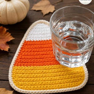 May include: A triangular crochet coaster shaped like a candy corn, featuring horizontal bands of white, orange, and yellow yarn with a cream-colored border. A clear glass of water sits on the coaster, which is positioned on a dark wooden surface.
