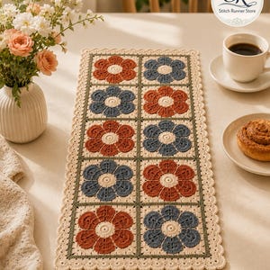 May include: Crocheted table runner featuring a repeating floral pattern in rust and blue, framed by a green border and a scalloped edge. The runner is placed on a light-colored surface, with a vase of flowers and a cup of coffee in the background.