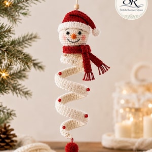 May include: A crocheted snowman ornament featuring a red hat, a red scarf, and a long, spiraling white body decorated with small red beads. The snowman has a round head with a small orange nose and a red pom-pom at the bottom of the spiral.