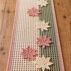 Pode incluir: Caminho de mesa retangular de crochê com centro creme, faixa verde e borda rosa. Folhas de bordo de crochê em tons de rosa, creme e verde espalhadas pelo caminho de mesa. Logotipo da Stitch Runner Store.