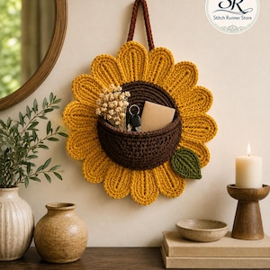 May include: A sunflower-shaped crochet wall hanging with a dark brown center pocket, yellow petals, and a green leaf. The pocket holds dried flowers, a key ring, and a small card. A brown loop at the top allows for hanging. Text reads Stitch Runner Store.