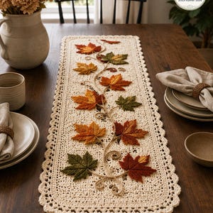 May include: A rectangular cream-colored crochet table runner featuring an intricate lace border and a central vine design adorned with three-dimensional fabric maple leaves in shades of orange, red, yellow, and green, arranged along the center.