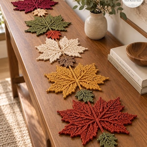May include: A crochet table runner featuring a series of interconnected maple leaves in various autumn colours including deep red, olive green, cream, brown, and golden yellow, arranged in a linear pattern along the length of the wooden furniture.