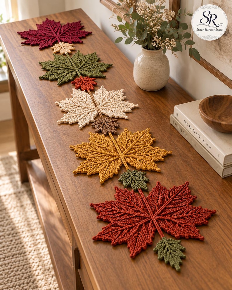 May include: A crochet table runner featuring a series of interconnected maple leaves in various autumn colours including deep red, olive green, cream, brown, and golden yellow, arranged in a linear pattern along the length of the wooden furniture.