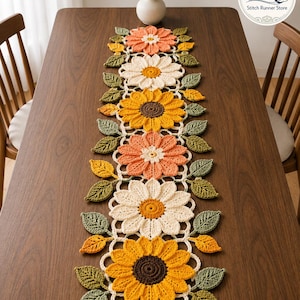 May include: A rectangular crochet table runner featuring a repeating pattern of large, multi-petaled flowers in shades of peach, cream, and yellow, accented with dark brown centers and surrounded by crocheted green and yellow leaves.