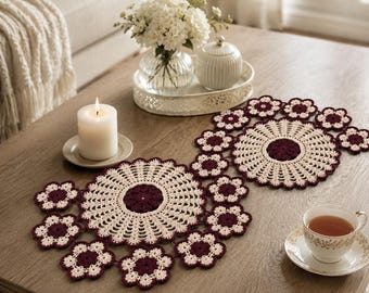 Könnte beinhalten: Zwei cremefarbene und bordeauxfarbene Häkeldeckchen mit floralem Design auf einem Holztisch. Eine brennende Kerze, eine Teetasse und eine Vase mit weißen Blumen sind ebenfalls vorhanden. Die Deckchen sind von kleineren blütenförmigen Häkelstücken umgeben.