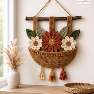 May include: A wall hanging featuring a brown crocheted basket suspended from a wooden branch. The basket contains three crocheted flowers in rust and cream, accented by green leaves. Three tassels in cream, gold, and rust hang from the bottom.