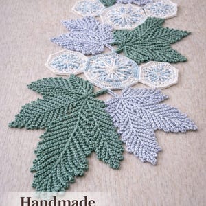 May include: Handmade crochet runner featuring leaf and circular designs in shades of green, blue, and white. The runner is made of yarn and is displayed on a neutral surface. The text "Handmade Crochet Runner" is visible.