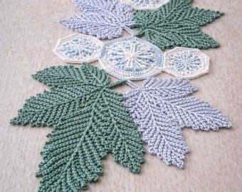 Könnte beinhalten: Handgefertigter Häkel-Tischläufer mit Blatt- und Kreisdesigns in Grün, Blau und Weiß. Der Läufer ist aus Garn gefertigt und auf einer neutralen Oberfläche platziert. Der Text "Handmade Crochet Runner" ist sichtbar.