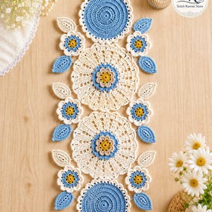 May include: A handmade crochet table runner with a floral design. The runner features cream-colored, blue, and yellow crochet flowers and leaves. The runner is placed on a light brown wooden surface.