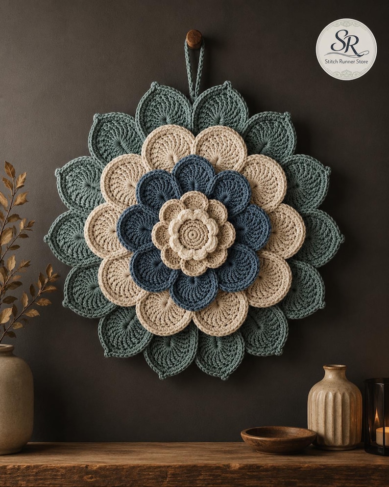 May include: A circular crochet wall hanging featuring a layered floral design with concentric rings of petals in shades of teal, cream, and dark blue. The piece includes a small hanging loop at the top and displays the text SR Stitch Runner Store.