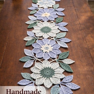 Könnte beinhalten: Handgefertigter gehäkelter Tischläufer mit floralem Design. Der Läufer zeigt blaue, grüne und weiße Blumen und Blätter. Der Text "Handmade Crochet Runner" ist sichtbar. Auf einem Holztisch.