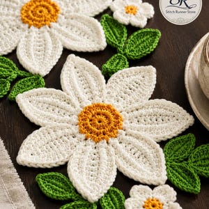 K&ouml;nnte beinhalten: Geh&auml;kelte Untersetzer und Dekorationen in G&auml;nsebl&uuml;mchenform in Wei&szlig; mit gelben Mitten und gr&uuml;nen Bl&auml;ttern. Das Set enth&auml;lt gro&szlig;e und kleine Blumenmotive. Ein kreisf&ouml;rmiges Logo in der oberen rechten Ecke zeigt den Text Stitch Runner Store.