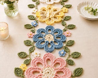 May include: A colorful, crocheted floral table runner. The runner features large, openwork flowers in shades of pink, white, yellow, and blue, connected by green vines with leaves and small flowers. A candle and vase of flowers are in the background.