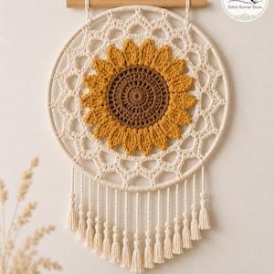 May include: A circular wall hanging featuring a crocheted sunflower with a brown center and yellow petals, set within a cream-colored macrame web on a wooden dowel. The piece includes a hanging cord and a row of cream-colored tassels at the bottom.