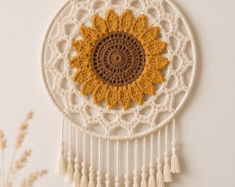 May include: A circular wall hanging featuring a crocheted sunflower with a brown center and yellow petals, set within a cream-colored macrame web on a wooden dowel. The piece includes a hanging cord and a row of cream-colored tassels at the bottom.