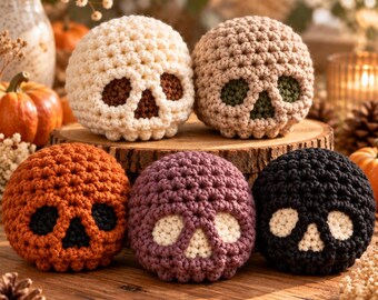 May include: Five spherical crochet skulls in cream, beige, orange, purple, and black. Each skull features a contrasting color for the eye sockets and nose cavity. The cream skull has brown details, the beige has green, the orange has black, and the purple and black skulls have cream-colored facial features.