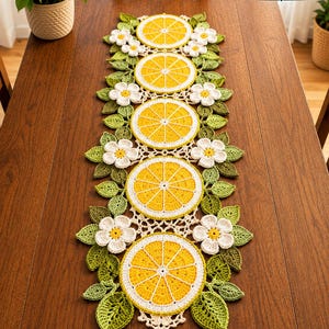 May include: A handmade crochet table runner featuring a repeating pattern of yellow lemon slices, white flowers, and green leaves. The runner is placed on a wooden table. The Stitch Runner Store logo is in the top right corner.