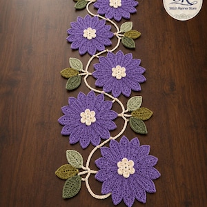 May include: A handmade crochet table runner in purple, with a floral pattern. The runner features large flowers with cream centres and green leaves. The design is set against a dark wood surface.