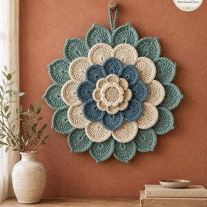 May include: A circular crochet wall hanging featuring layered, petal-like shapes in shades of teal, cream, and blue. The piece has a central cream flower, surrounded by concentric rings of blue and cream petals, finished with a teal outer border.