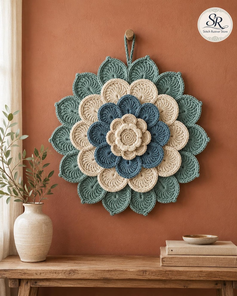 May include: A circular crochet wall hanging featuring layered, petal-like shapes in shades of teal, cream, and blue. The piece has a central cream flower, surrounded by concentric rings of blue and cream petals, finished with a teal outer border.