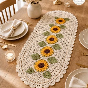 Puede incluir: Camino de mesa ovalado de ganchillo color crema con borde festoneado. Presenta un patrón repetido de girasoles amarillos y marrones con hojas verdes. El camino de mesa está sobre una mesa de madera.
