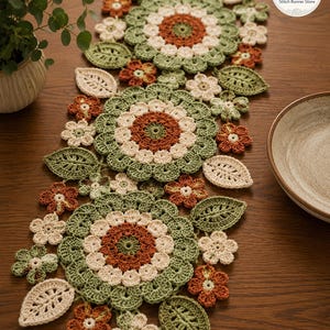 Può includere: Un runner da tavola all'uncinetto con un motivo ripetuto di grandi fiori circolari nei colori verde salvia, crema e arancione ruggine, intervallati da fiori più piccoli e foglie dettagliate nelle stesse tonalità.