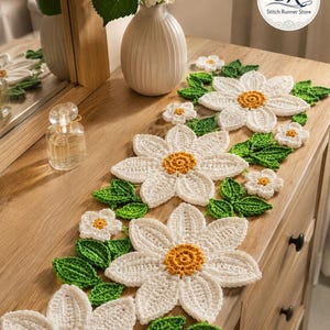 K&ouml;nnte beinhalten: Ein geh&auml;kelter Tischl&auml;ufer mit wei&szlig;en G&auml;nsebl&uuml;mchenmotiven, gelben Mitten und gr&uuml;nen Bl&auml;ttern. Das florale Muster ist in einer sich wiederholenden Abfolge entlang des L&auml;ufers angeordnet und zeigt detaillierte Stiche und strukturierte Bl&uuml;tenbl&auml;tter.