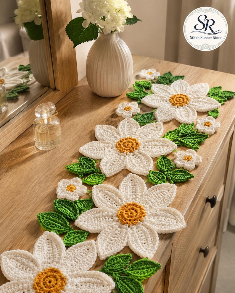 K&ouml;nnte beinhalten: Ein geh&auml;kelter Tischl&auml;ufer mit wei&szlig;en G&auml;nsebl&uuml;mchenmotiven, gelben Mitten und gr&uuml;nen Bl&auml;ttern. Das florale Muster ist in einer sich wiederholenden Abfolge entlang des L&auml;ufers angeordnet und zeigt detaillierte Stiche und strukturierte Bl&uuml;tenbl&auml;tter.