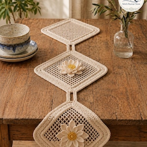 Op de afbeelding: Crèmekleurige, gehaakte tafelloper met een ruitpatroon en drie bloemenapplicaties. De loper wordt getoond op een houten tafel, die de textuur en het ontwerp laat zien. De bloemen hebben gele centra.
