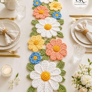 Könnte beinhalten: Gehäkelter Tischläufer mit Gänseblümchenmotiven in Weiß, Gelb, Orange und Blau, mit grünen Blättern. Der Läufer liegt auf einer weißen Tischdecke, mit Gedecken und Blumengestecken auf beiden Seiten.