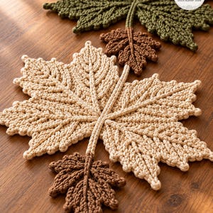 May include: A collection of crocheted leaf-shaped coasters in olive green, beige, and brown. The set includes one large olive green leaf, one large beige leaf, and two smaller brown leaves, all featuring detailed raised stitching and stems.