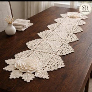 May include: Cream-colored, crocheted table runner with a floral design. The runner is placed on a dark brown wooden table. The runner features a repeating geometric pattern and decorative floral accents at each end.