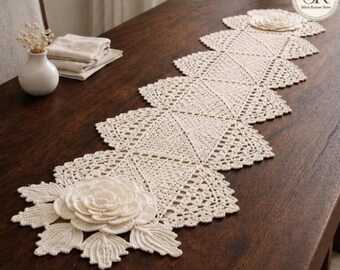 Könnte beinhalten: Cremefarbener, gehäkelter Tischläufer mit floralem Design. Der Läufer liegt auf einem dunkelbraunen Holztisch. Er weist ein sich wiederholendes geometrisches Muster und dekorative florale Akzente auf.