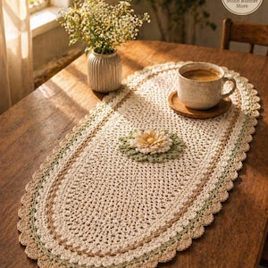 May include: Oval, cream-colored crochet table runner with a scalloped edge. The runner features a central floral design and is placed on a wooden table. A cup of coffee sits on a wooden coaster. A vase of flowers is also on the table.