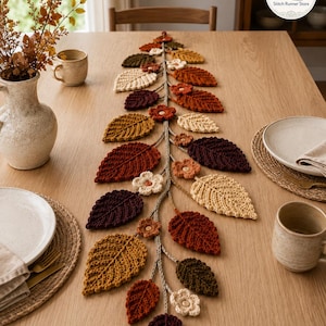 May include: A crochet table runner featuring a central vine with attached autumn-colored leaves in shades of brown, orange, yellow, and cream, interspersed with small crocheted flowers. The runner is displayed on a wooden table with place settings.