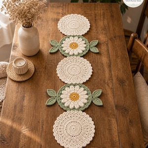 Puede incluir: Un conjunto de posavasos y un camino de mesa hechos a mano con ganchillo. Los posavasos son de color crema con un diseño circular. El camino de mesa presenta un diseño de margarita con hojas verdes y un centro amarillo. Se muestra sobre una mesa de madera.