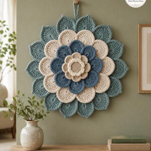 May include: A circular crochet wall hanging featuring layered, petal-like shapes in alternating shades of teal, cream, and blue. The design radiates from a central cream-colored flower, with a small loop at the top for hanging. The text SR Stitch Runner Store is visible in the upper right corner.