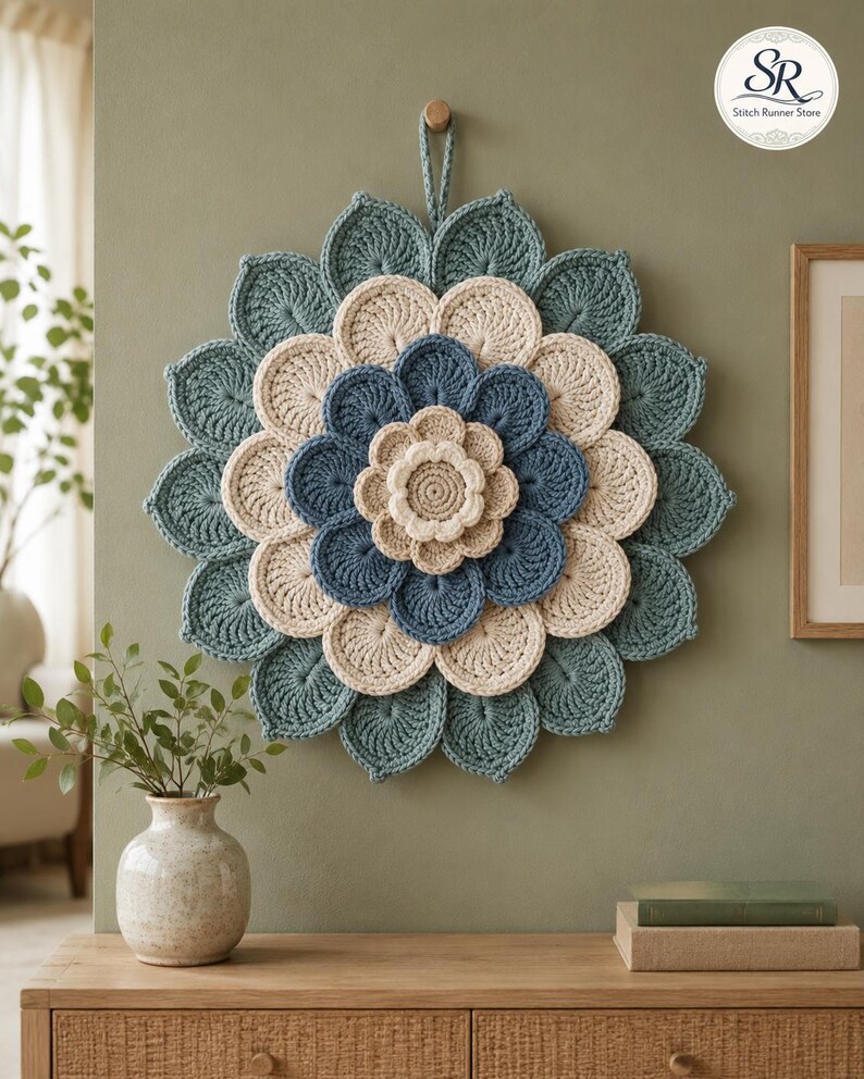May include: A circular crochet wall hanging featuring layered, petal-like shapes in alternating shades of teal, cream, and blue. The design radiates from a central cream-colored flower, with a small loop at the top for hanging. The text SR Stitch Runner Store is visible in the upper right corner.
