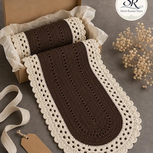 May include: A dark brown crochet table runner with a cream-colored scalloped lace border. The runner is partially folded inside a cardboard gift box lined with tissue paper, with a portion extending outward. A blank brown paper tag is visible.