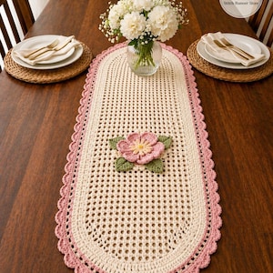 May include: Cream-coloured, oval crochet table runner with a pink scalloped border. A pink and yellow flower with green leaves is centered on the runner. The runner is on a dark wooden table, with plates and flowers.