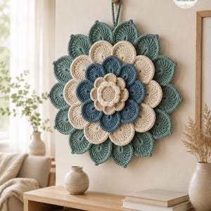 May include: A circular crochet wall hanging featuring layered, petal-like shapes in shades of sage green, cream, and dusty blue. The design is arranged in a concentric floral pattern with a hanging loop at the top and a Stitch Runner Store logo.