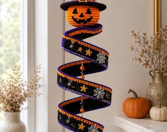 Könnte beinhalten: Eine hängende Häkeldekoration bestehend aus einem Kürbiskopf mit schwarzem Hexenhut, befestigt an einem spiralförmigen schwarzen Band mit orangefarbenem und violettem Rand, verziert mit gelben Sternen, weißen Spinnennetzen und zwei kleinen schwarzen Spinnen.