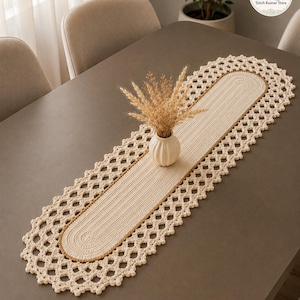 May include: An oval-shaped cream-coloured crochet table runner featuring a central ribbed texture, a thin tan border, and an intricate openwork scalloped edge. A small white ribbed vase containing dried wheat stalks sits in the centre of the runner.