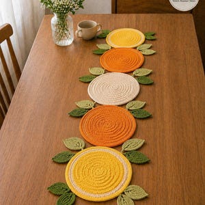 Könnte beinhalten: Handgefertigter Tischläufer mit Zitrusfrucht-Design. Der Läufer hat kreisförmige Tischsets in Gelb, Orange und Creme, jeweils mit grünen Blättern. Die Tischsets sind auf einem Holztisch in einer Reihe angeordnet.