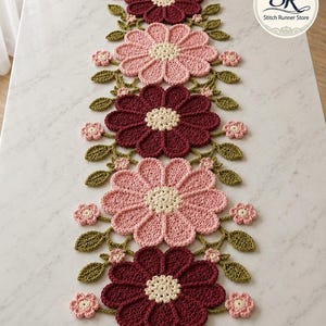 May include: A crochet table runner with a floral pattern. The runner displays large flowers in pink and burgundy, with cream centres and green leaves. The text "Vintage Floral Runner" is visible.